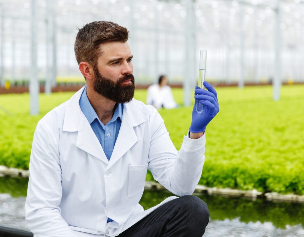 Thoughtful man researcher holds a glass tube with sample standing before plants in the greenhouse